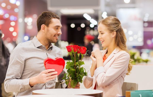 love, romance, valentines day, couple and people concept - happy young man with red flowers giving present to smiling woman at cafe in mall